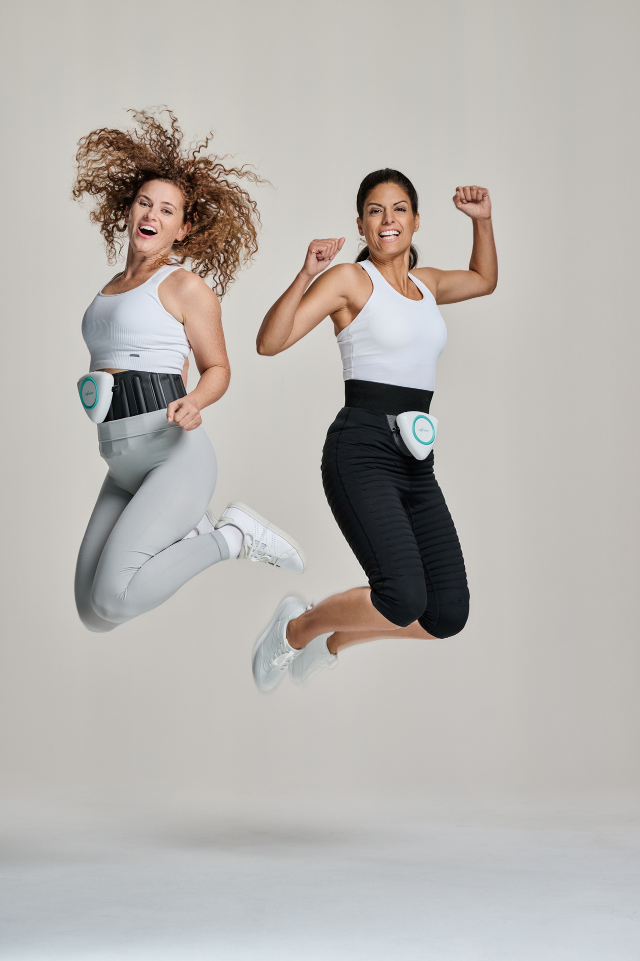 Zwei Frauen springen fröhlich mit Fitness-Gürteln an der Taille, tragen Sportkleidung und posieren vor hellem Hintergrund.
